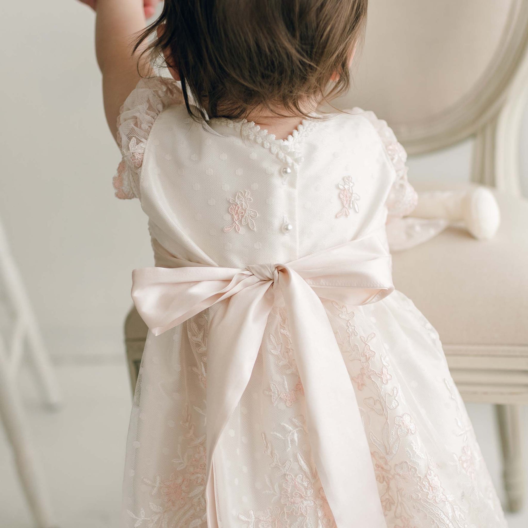 A young child is seen from behind, wearing the Elizabeth Christening Dress, a handmade ivory gown adorned with lace and floral embroidery. This elegant silk Dupioni dress features a large satin bow elegantly tied at the back and delicate pearl buttons. The child has dark hair and is reaching up, possibly holding onto something out of frame.