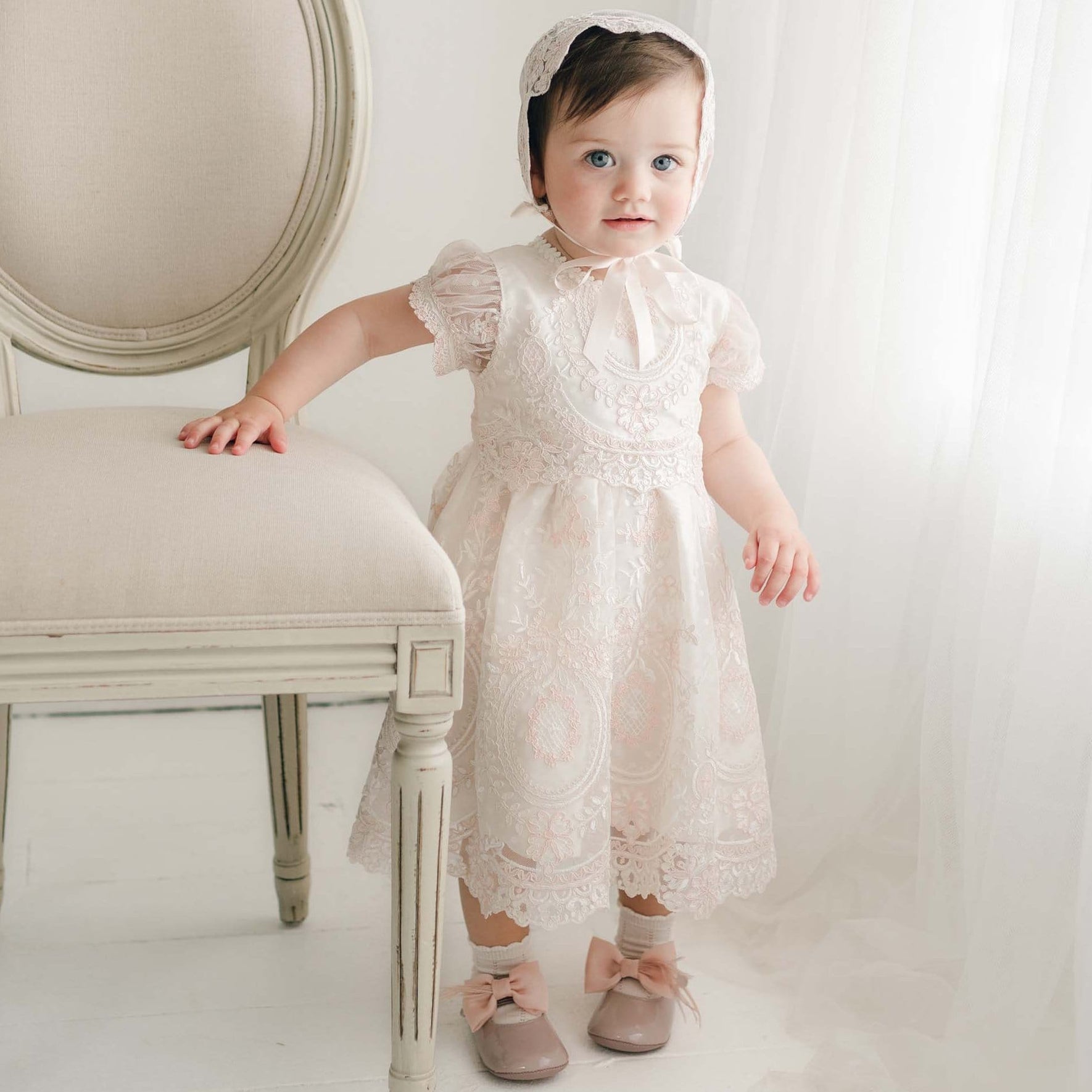A young child stands next to a white chair in a softly lit room, wearing the exquisite Elizabeth Christening Dress and matching bonnet, paired with pink shoes adorned with bows. One hand rests on the chair while the other hangs by their side. The delicate Christening dress enhances the scene, accompanied by light curtains in the background.
