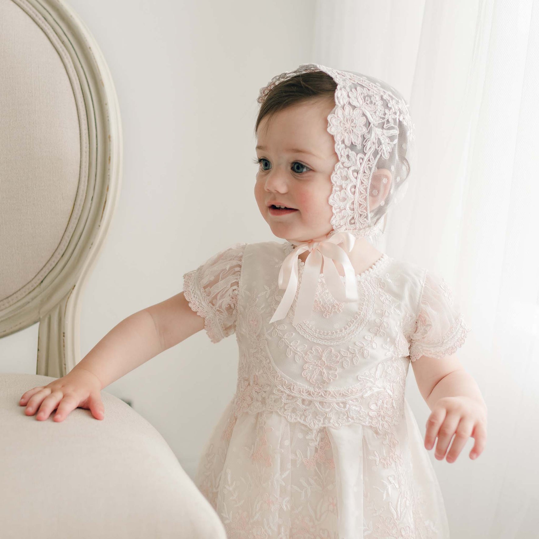 A young child stands by an upholstered chair, wearing the Elizabeth Christening Dress and a handmade white lace bonnet with a pink ribbon. The child has an attentive expression with a subtle smile, in a softly lit room with white curtains.