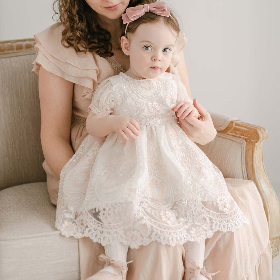 A woman with curly hair sits on a beige upholstered chair, holding a young girl on her lap. The girl is wearing the Elizabeth Christening Dress, featuring lace details and accessorized with a pink bow in her hair and matching pink shoes. Both look at something out of the frame with neutral expressions.