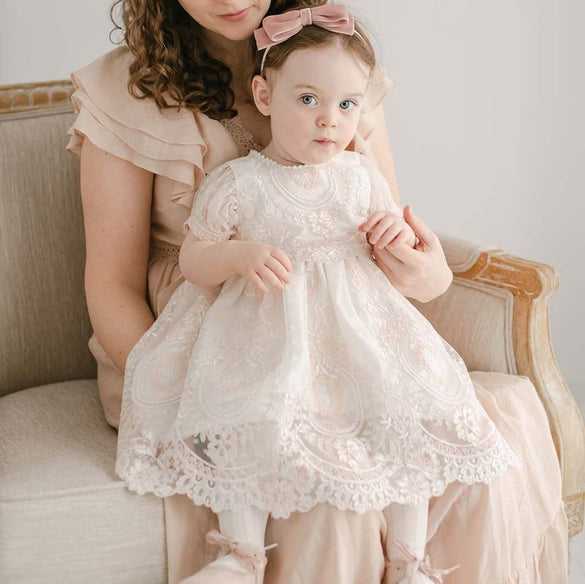 A woman with curly hair sits on a beige upholstered chair, holding a young girl on her lap. The girl is wearing the Elizabeth Christening Dress, featuring lace details and accessorized with a pink bow in her hair and matching pink shoes. Both look at something out of the frame with neutral expressions.