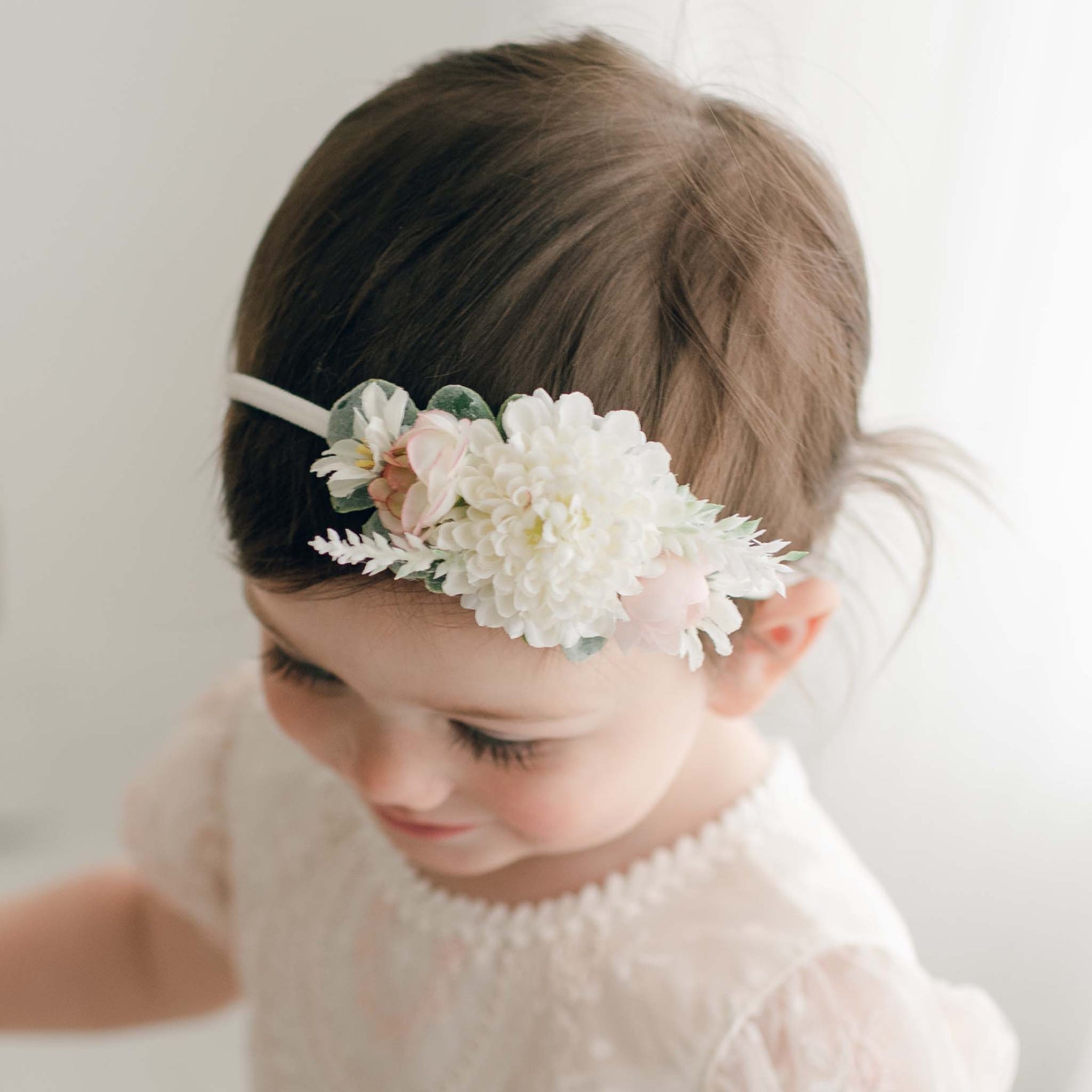A young child with brown hair is wearing the Elizabeth Flower Headband and a light-colored lace dress. The child appears to be looking down, with long eyelashes visible against a plain, softly lit background that exudes an aura of baby comfort.