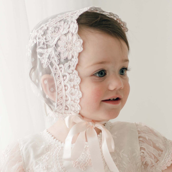Side view of a baby girl wearing the Elizabeth Christening Bonnet, adorned with intricate floral lace and tied with silk ribbons beneath her chin, and matching gown.
