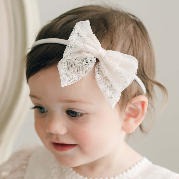 A young child with brown hair is wearing the Elizabeth Lace Bow Headband and looking slightly down and to the side, with a faint smile. The background is neutral, and the child is dressed in a white outfit featuring delicate detailing.