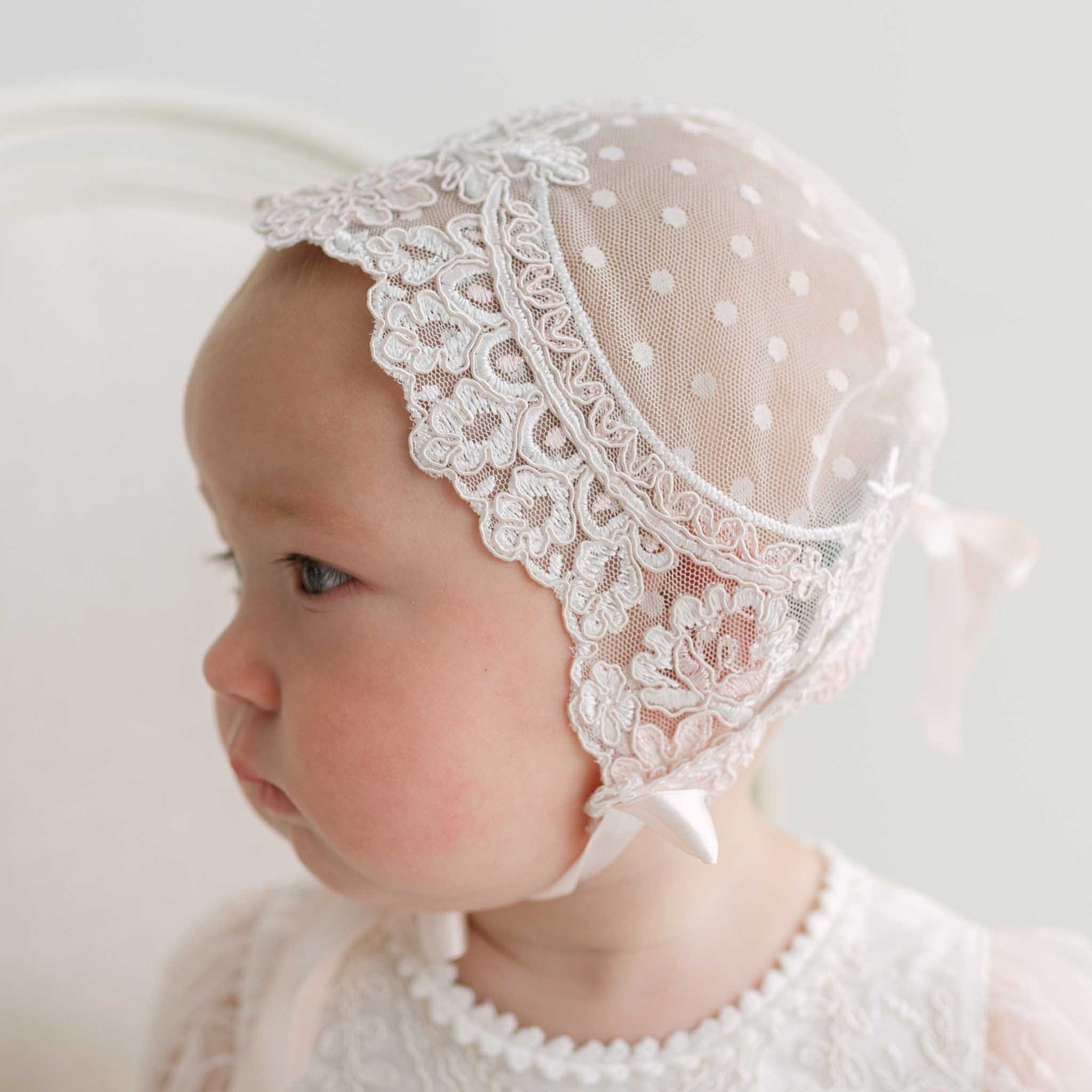 A baby wearing an ivory and pink lace bonnet with ivory polka-dot mesh and a chin strap, shown in profile against a soft white background.