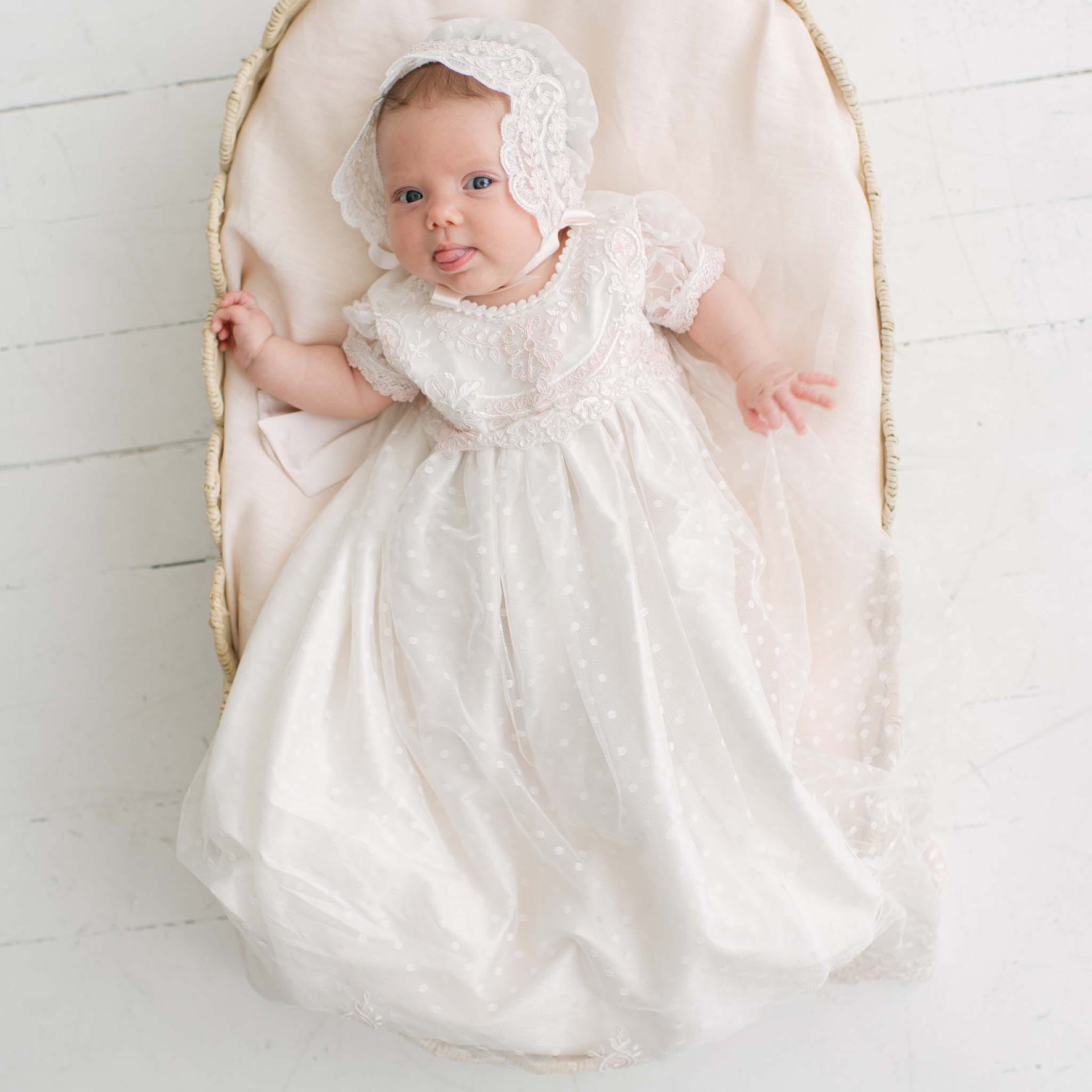 A baby wearing an ivory and pink lace and tulle blessing dress with a matching bonnet, lying on a cream-colored blanket inside a wooden bassinet.