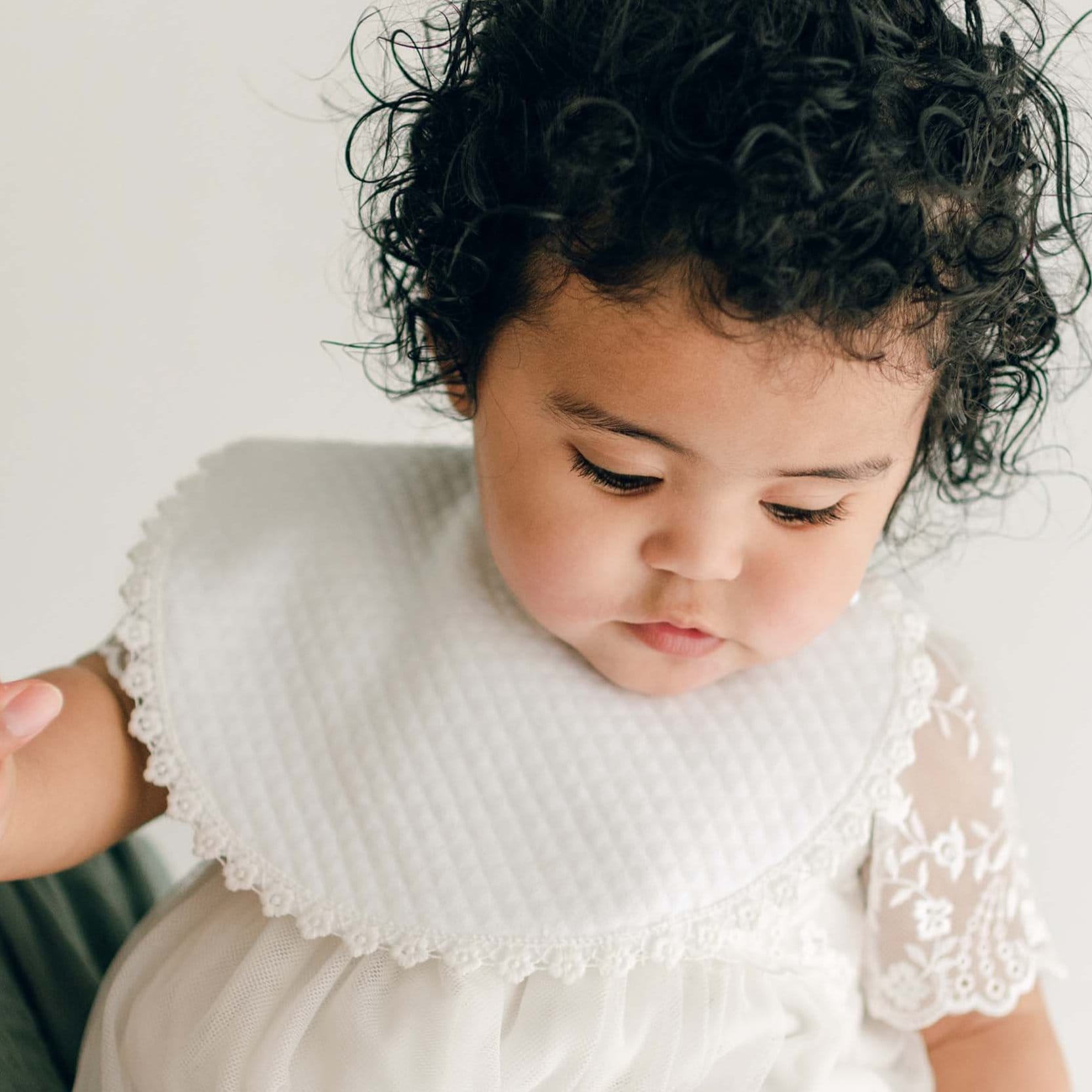 A baby with curly black hair, wearing a white baptism outfit and an Ella Bib with lace trim, is looking down.