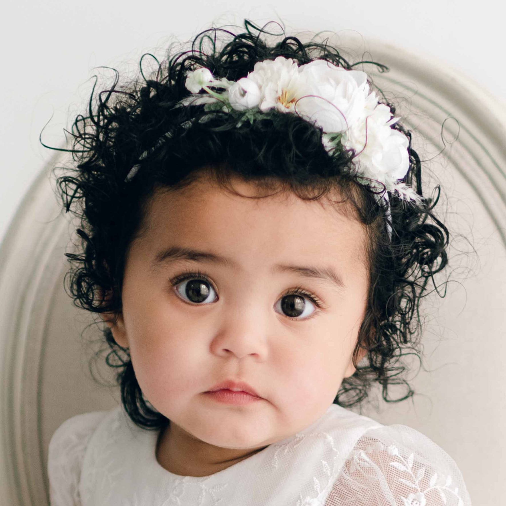 A baby girl with curly dark hair adorned with the Ella Flower Headband, featuring white flowers, looks directly at the camera.