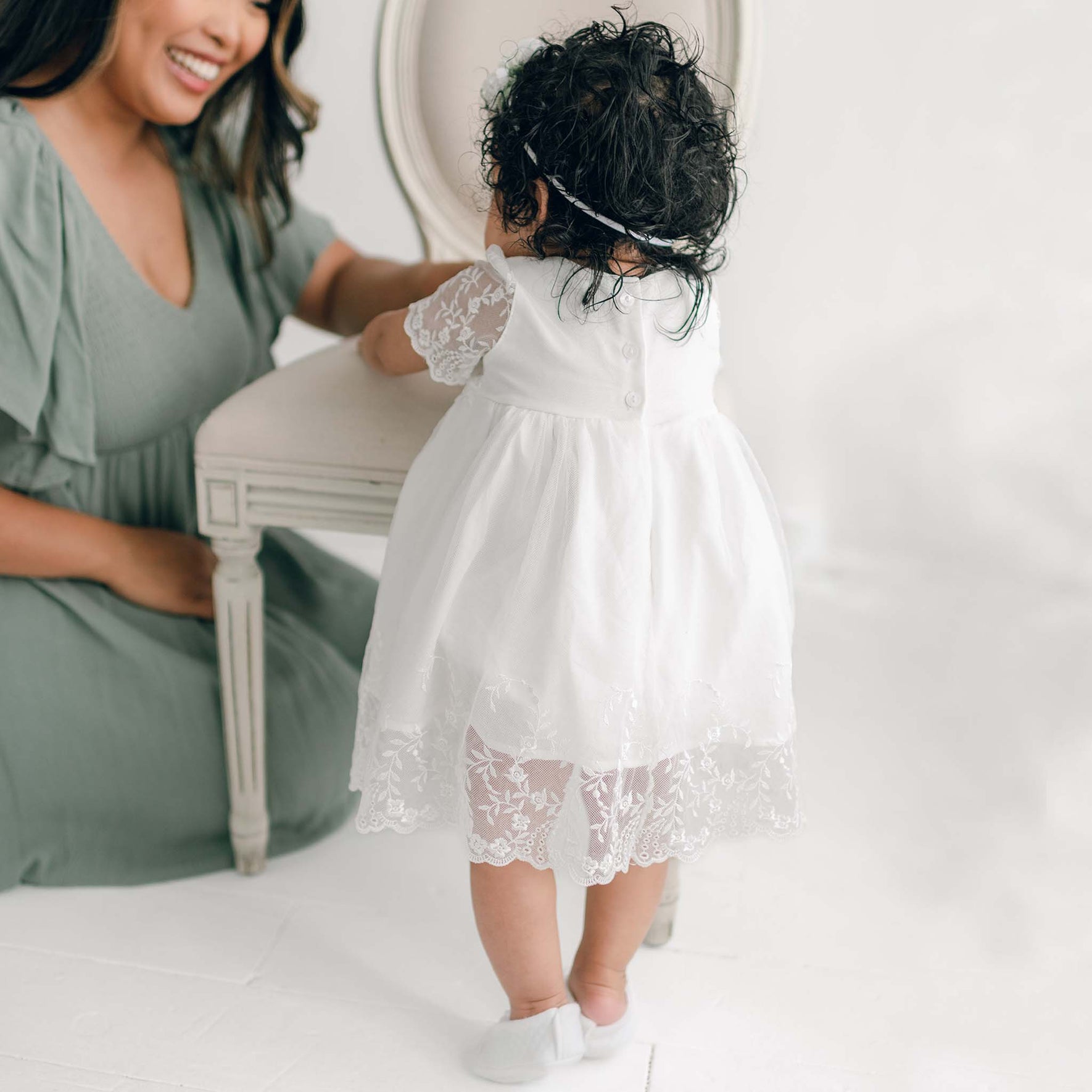 A woman in a light green dress kneels next to her baby daughter wearing an Ella Romper Dress.