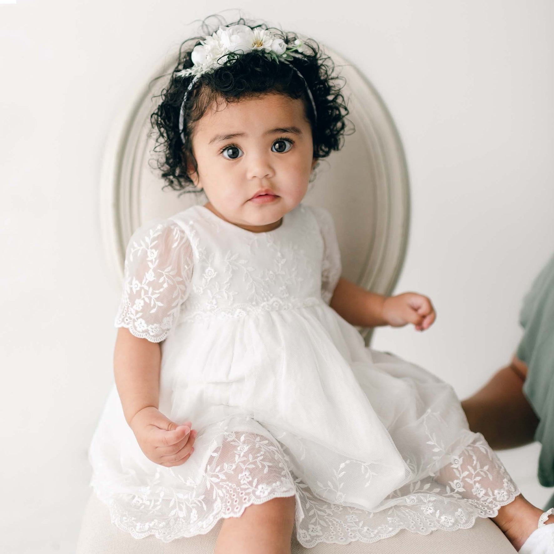 A baby girl with curly hair, wearing the Ella baptism dress flower headband, sits on an light beige chair and looks forward with a curious expression.