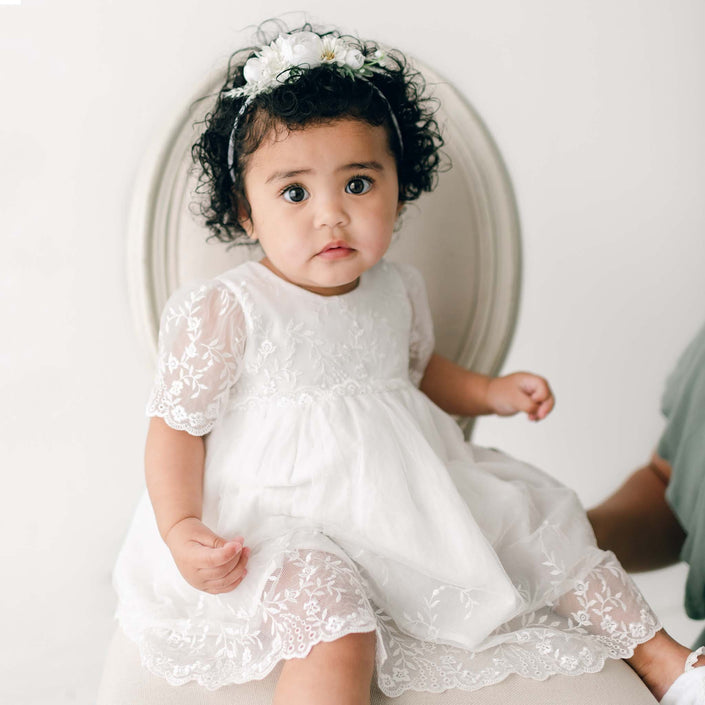A baby girl with curly hair, wearing the Ella baptism dress flower headband, sits on an light beige chair and looks forward with a curious expression.