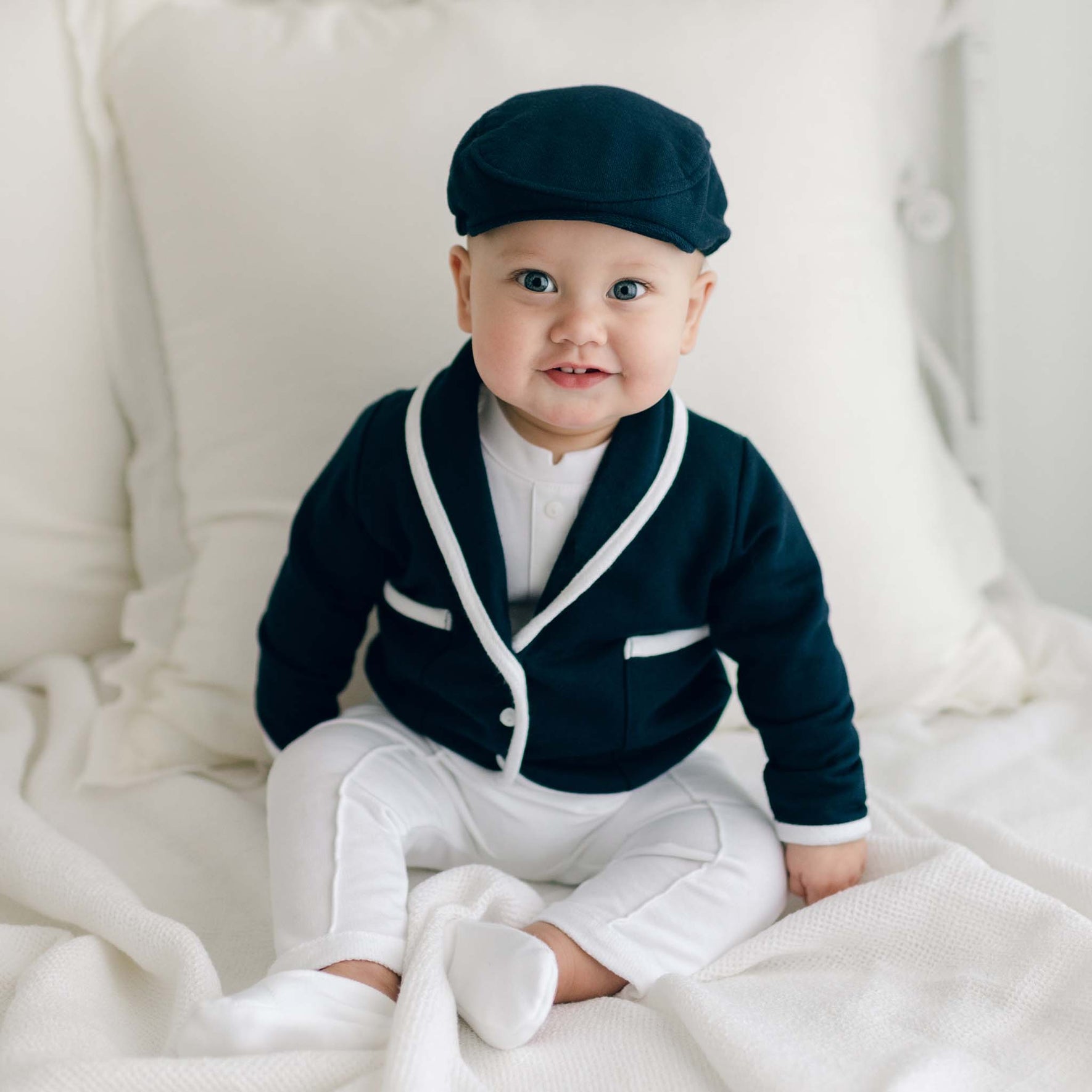 Smiling baby boy sitting on a bed and wearing the Elliott 3-Piece Suit, including the jacket, pants, and onesie (and matching newsboy cap).