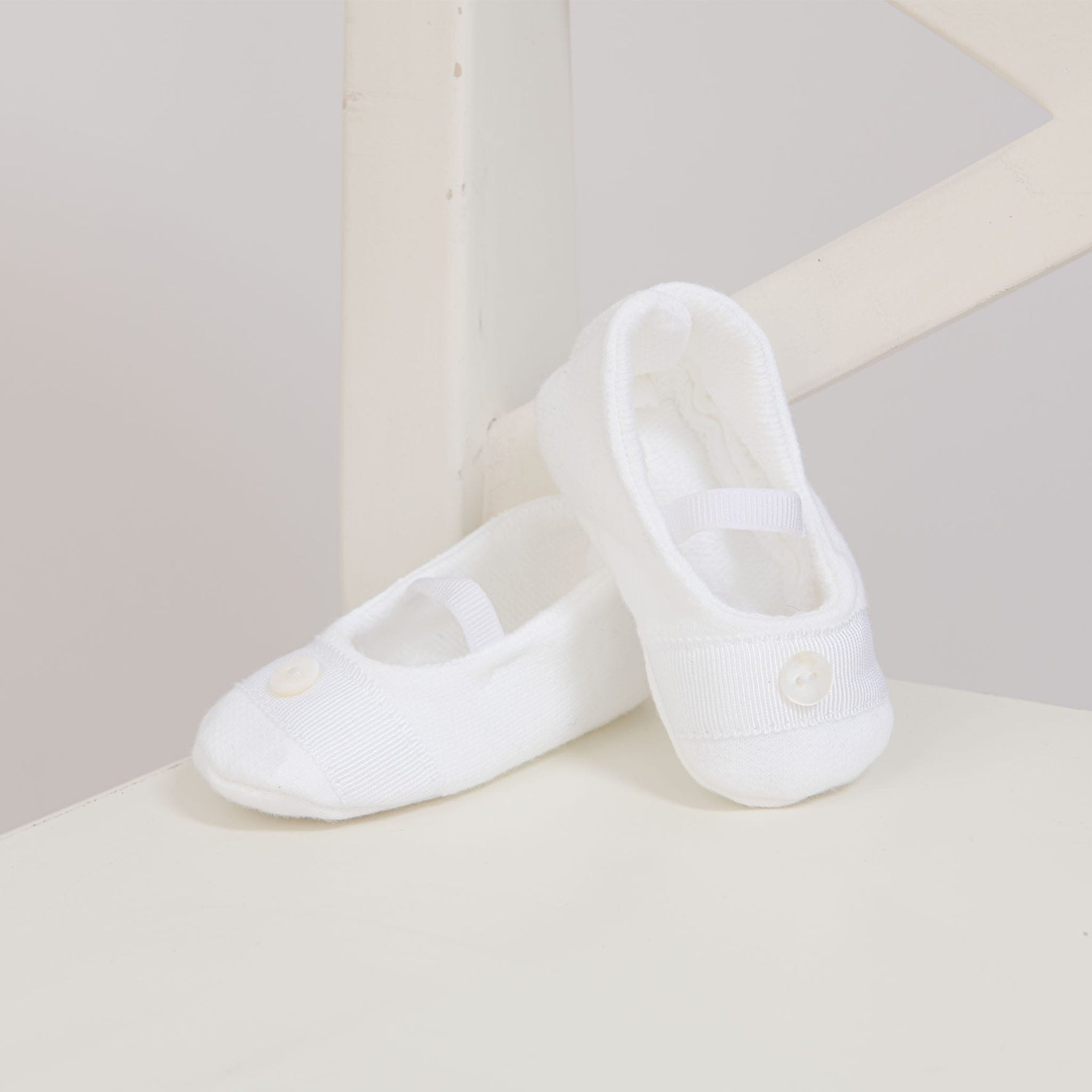 The Elliott Booties, a pair of small white baby booties featuring a ribbed texture and button detail, are perfect for Christening Day. They rest against a white wooden chair, showcasing elastic straps across the top. Their simplicity and charm are highlighted by the neutral background.