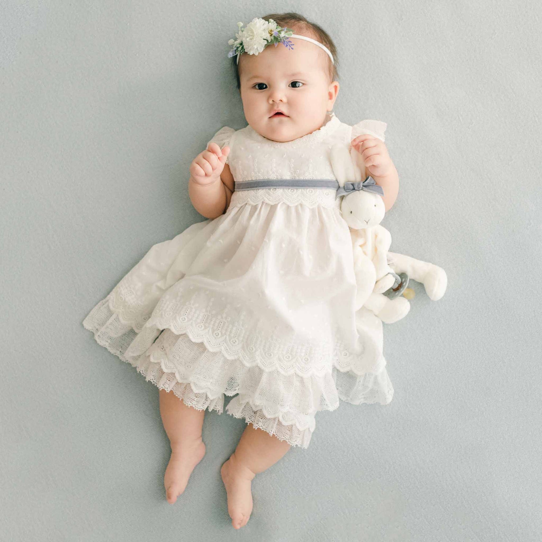 A baby lying on a soft blue surface in the Emily Dress & Bloomers, accessorized with a floral headband and a coordinating plush bunny.