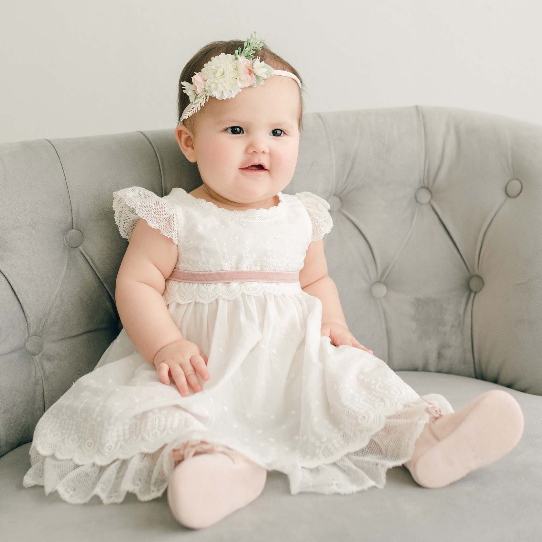 A baby in the Emily Dress & Bloomers, styled with a pink velvet ribbon and matching blush shoes, sitting on a gray sofa.