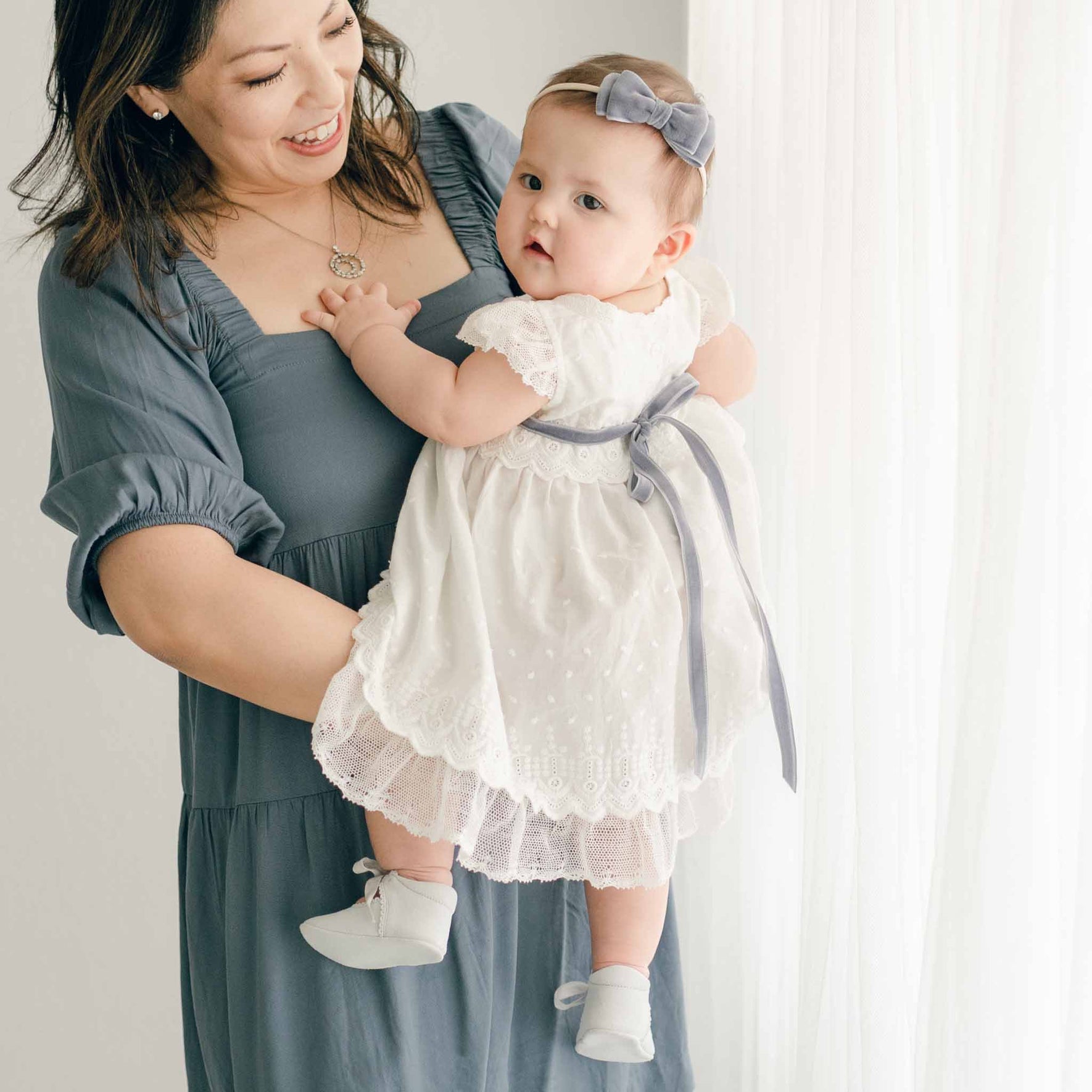 A mother holding her baby dressed in the Emily Dress & Bloomers, featuring a soft gray velvet ribbon tied at the back.