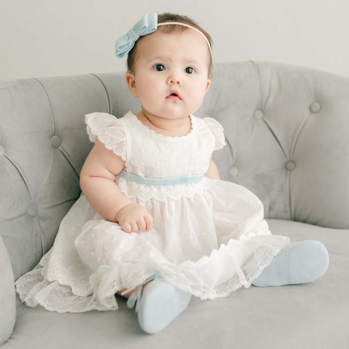 A baby in the Emily Dress & Bloomers sits on a tufted gray sofa, wearing a pastel blue velvet ribbon and matching shoes.