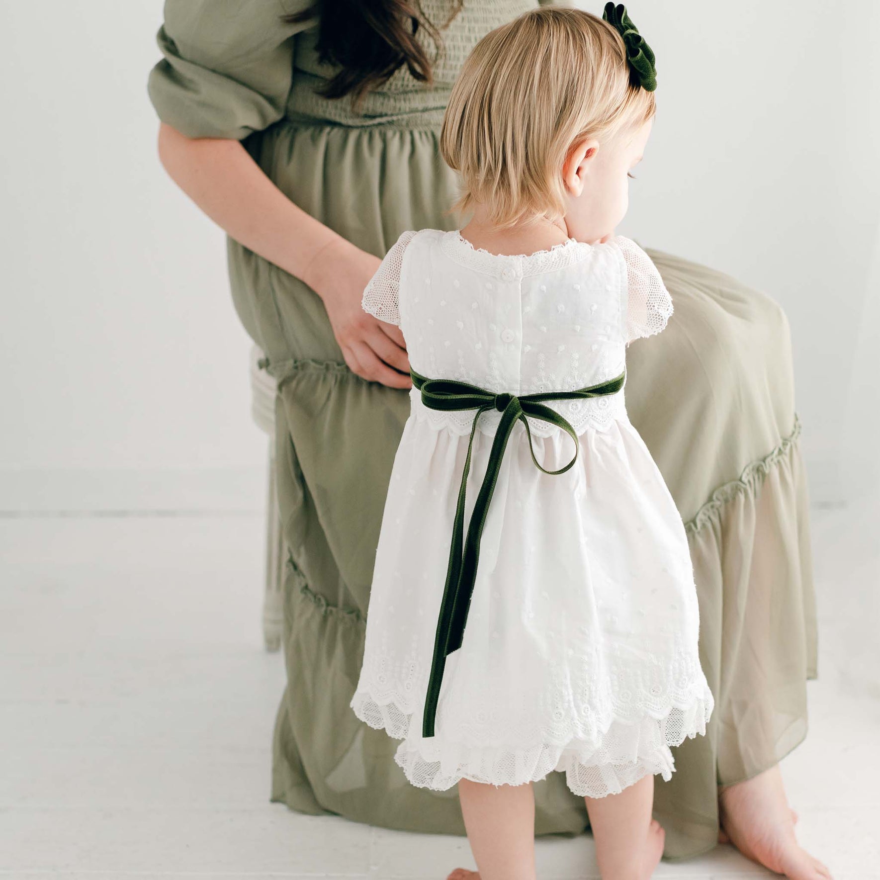 A woman in a green dress sits as a small child, facing away, wears the Emily Dress & Bloomers adorned with a green sash. The softly lit background provides a calm and gentle atmosphere, ideal for creating a timeless Christmas look.