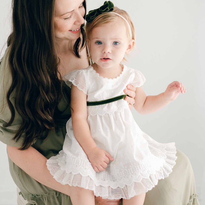 A woman with long hair, wearing a green outfit reminiscent of Christmas, smiles while holding a young child. The child is dressed in the Emily Dress & Bloomers, featuring lace details and a green velvet ribbon sash, and happily sits with her against a light-colored background.