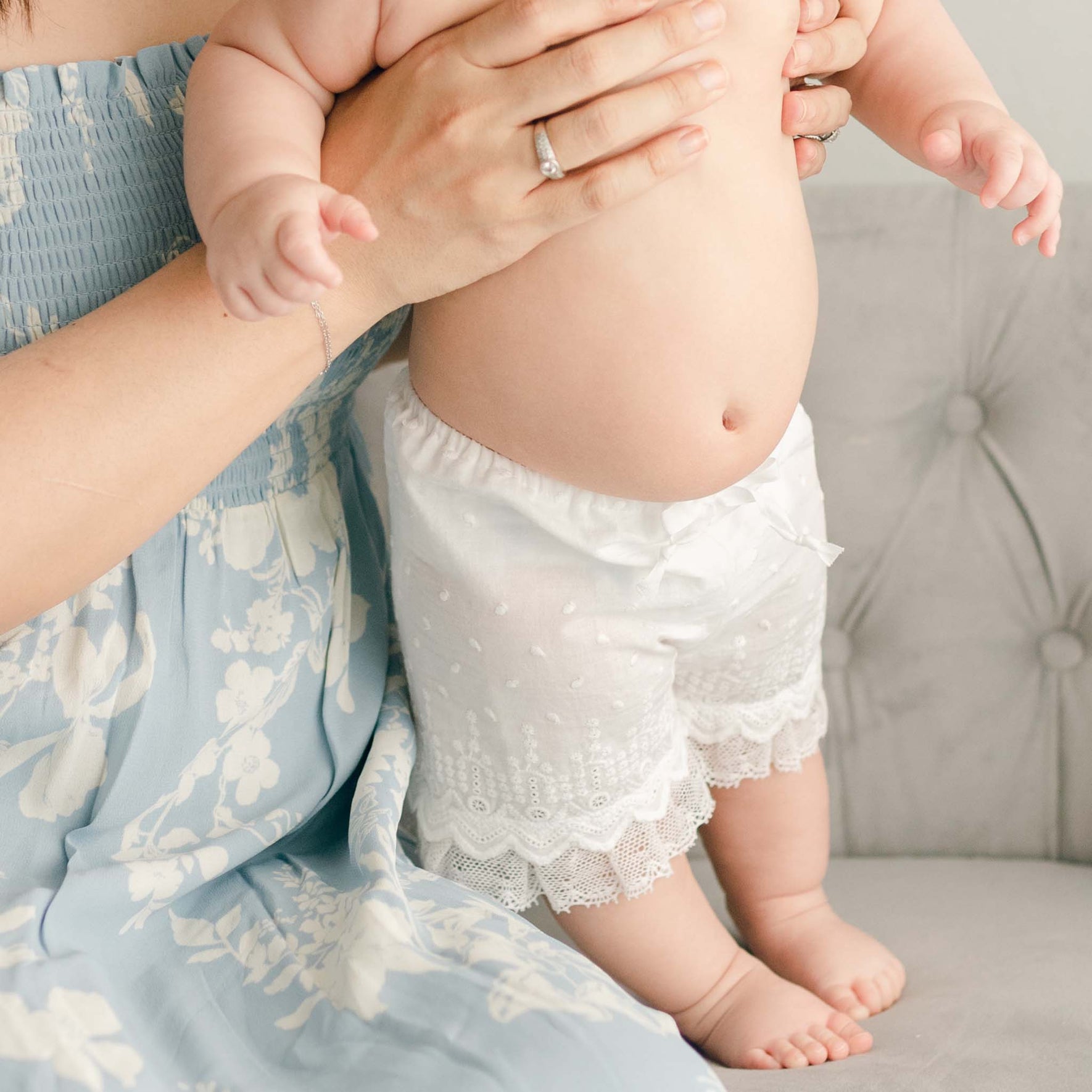 A woman cradles a baby in embroidered cotton lace shorts, perfectly complementing her own blue floral Emily Dress & Bloomers with a Red Sash. She wears a delicate ring on her finger, with the scene set against a light gray, tufted sofa.