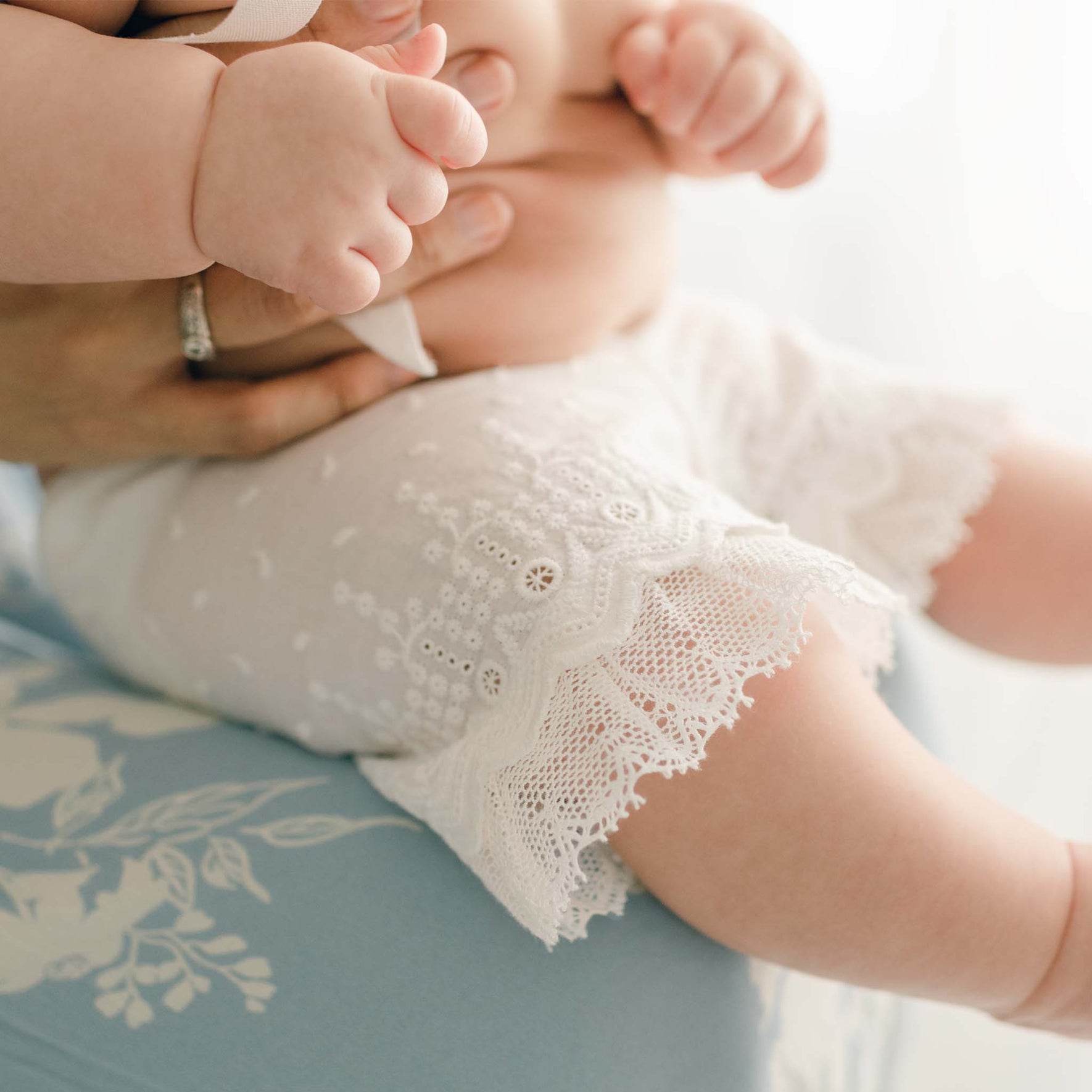 A baby wearing white lace-trimmed shorts, reminiscent of the Emily Dress & Bloomers with a Red Sash, is cradled gently on an adult's lap. The adult wears a light blue garment, which beautifully accentuates the delicate embroidered cotton lace on the baby's shorts and tiny hands.