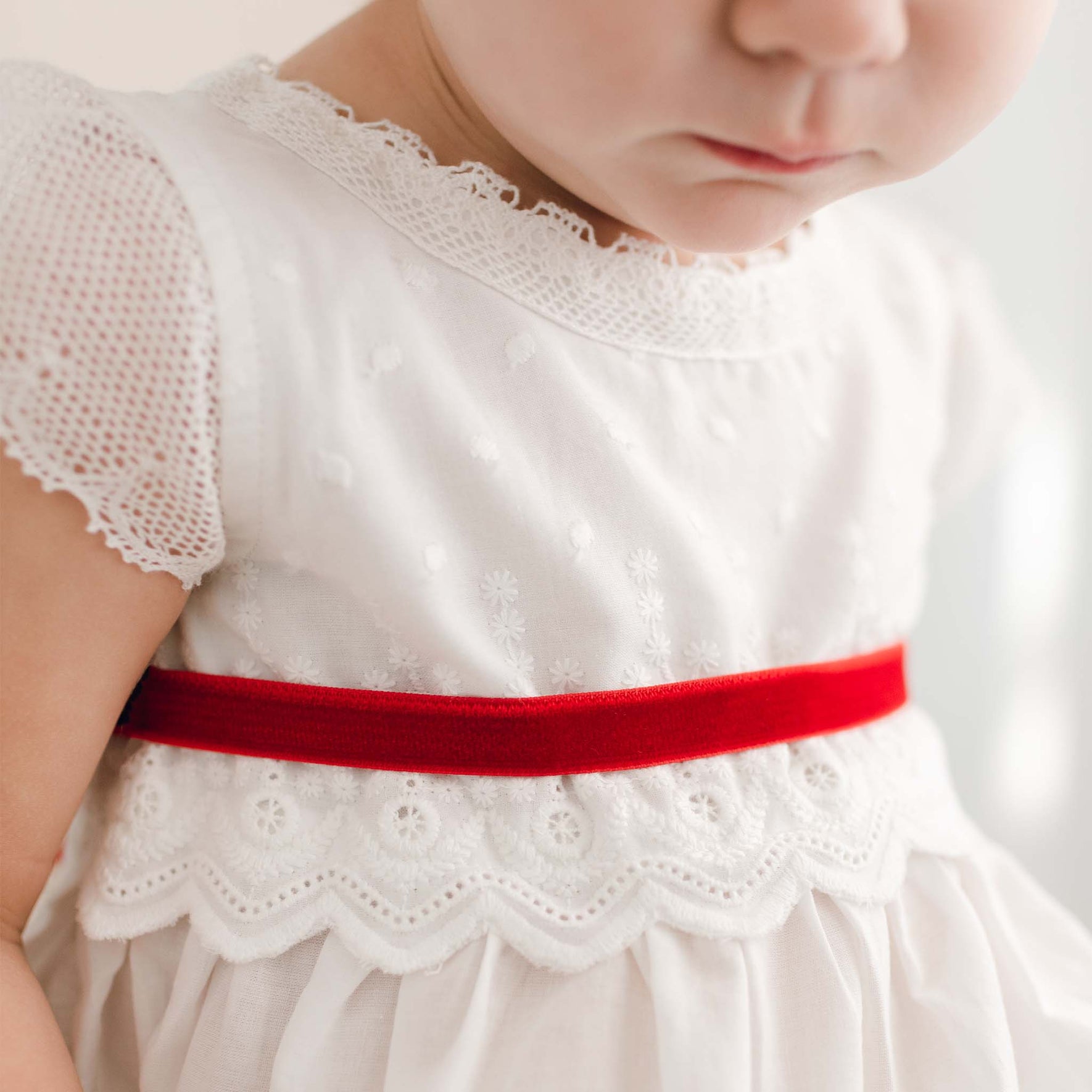 A young child is wearing the Emily Dress & Bloomers with a Red Sash, an exquisite Christmas outfit adorned with embroidered cotton lace and a vibrant red waistband. The dress features textured fabric patterns and a scalloped edge, highlighting its intricate details even though the child is only partially visible.