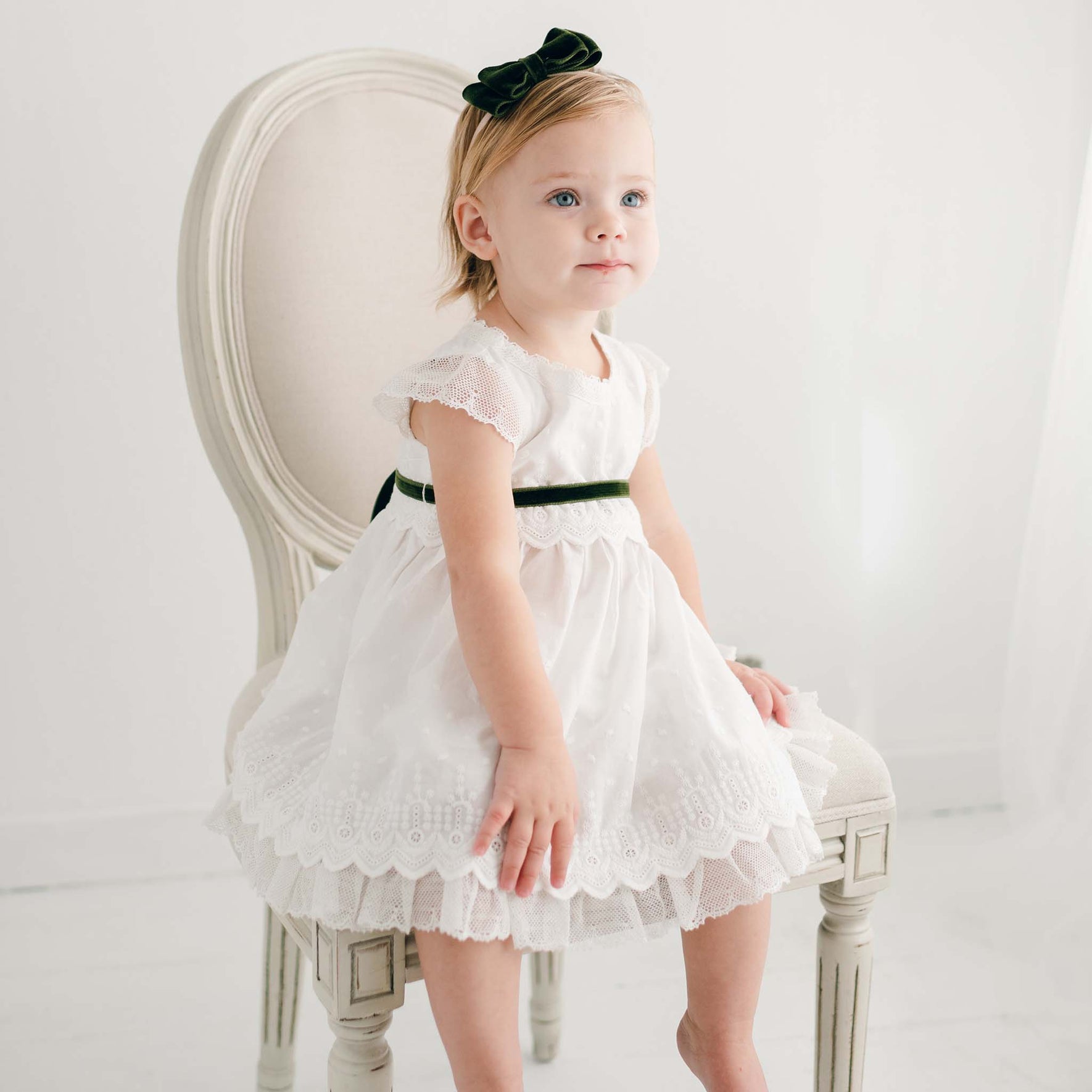 A toddler in a white dress with lace details and a black belt is sitting on a white upholstered chair, adorned with the Emily Green Velvet Bow Headband in their hair, looking slightly upward. The setting is bright with a neutral background.