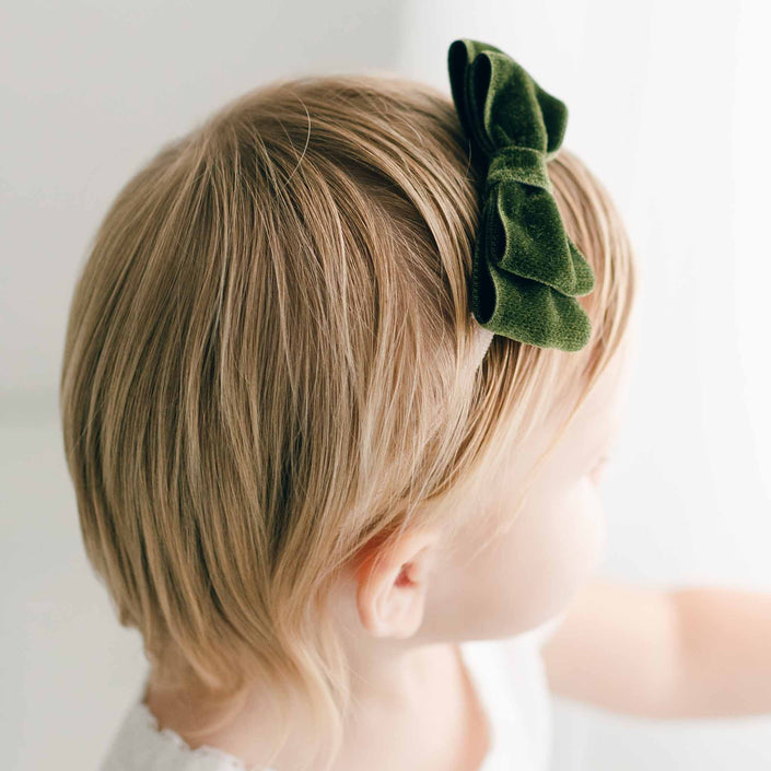 A young child with short blonde hair is wearing an Emily Green Velvet Bow Headband, handmade in the USA, facing away from the camera against a softly lit background.