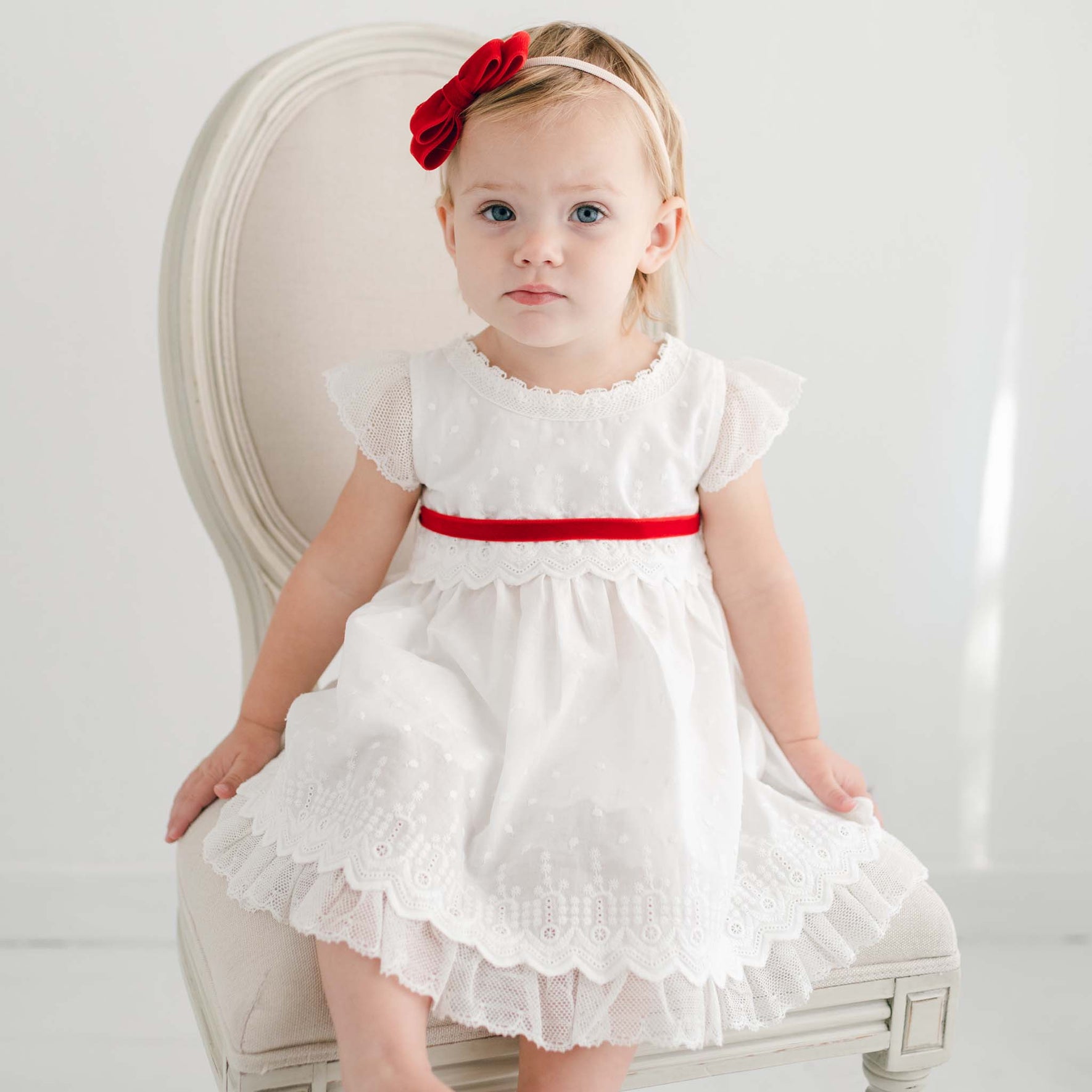 A toddler in a white dress adorned with lace details and a red ribbon sits on a cream-colored chair. She wears the Emily Red Velvet Bow Headband, handmade in the USA, as she gazes toward the camera against a plain, light-colored wall.
