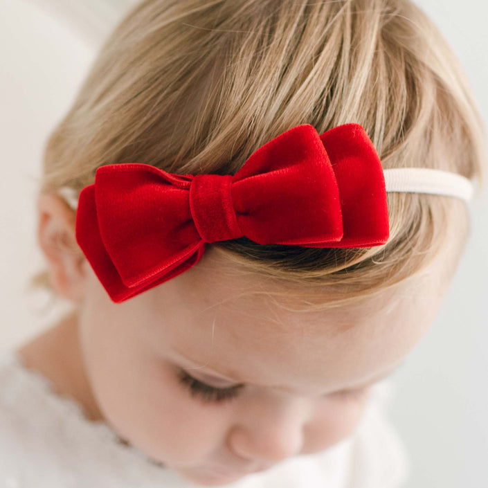 A young child with light hair wears the Emily Red Velvet Bow Headband, featuring a super soft ribbon and a large red velvet bow. The child is facing downward, and the softly blurred background enhances the charming scene.