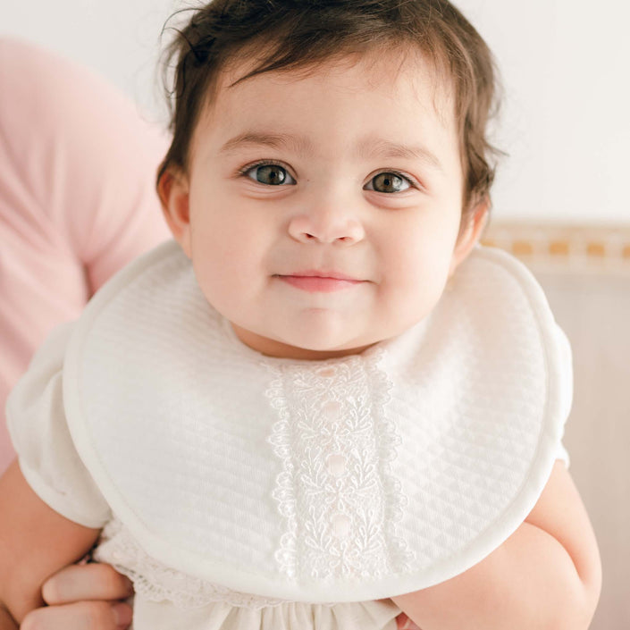 A baby with brown hair and an Emma Bib sits on a person's lap, smiling softly at the camera. The background is softly blurred, adorned with hints of neutral-toned home decor and an ivory quilted cotton throw that adds a touch of cozy elegance.