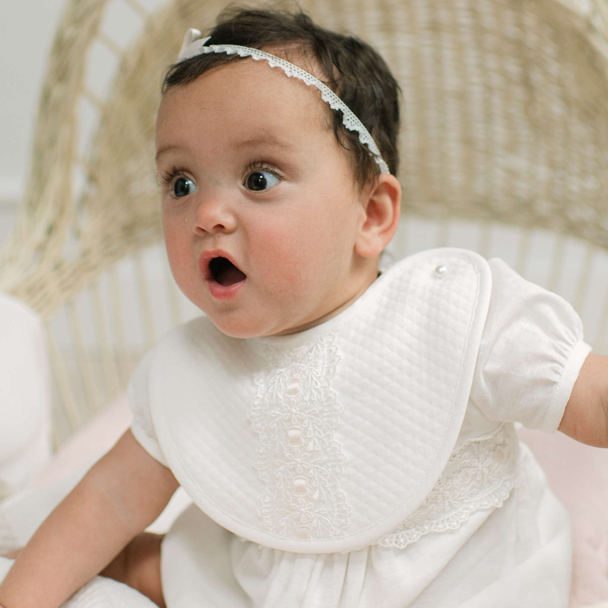 A baby wearing an ivory romper and a matching ivory quilted bib with lace trim, sitting in a woven chair. The baby also wears an ivory ace headband.