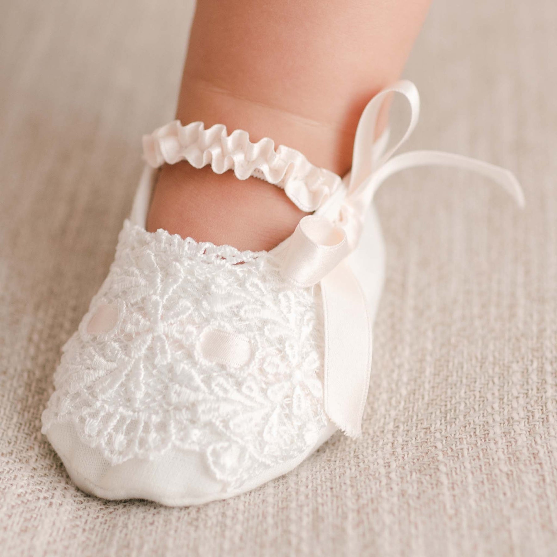 An infant's foot dressed in the Emma Christening Booties, featuring ivory lace and a pink silk ribbon bow, rests on a textured beige surface.