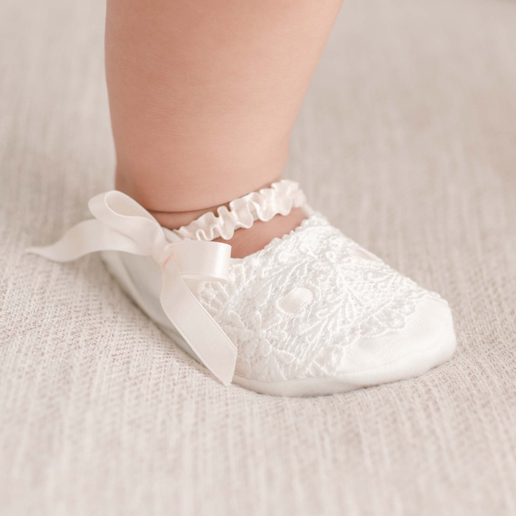A close-up of a baby's foot adorned in the Emma Christening Booties, featuring delicate white lace with pink silk ribbons. The baby stands on a soft, light-colored textured surface.