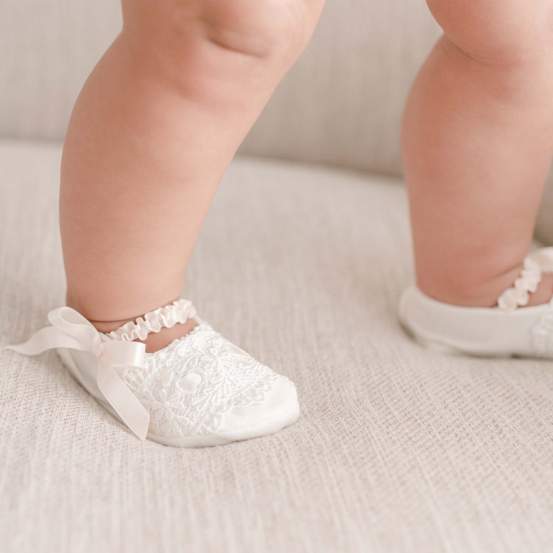 Close-up of a baby wearing the Emma Christening Booties, featuring ivory lace and adorned with pink silk ribbons, sitting on a light-colored fabric surface. The focus is on the baby's feet and booties.