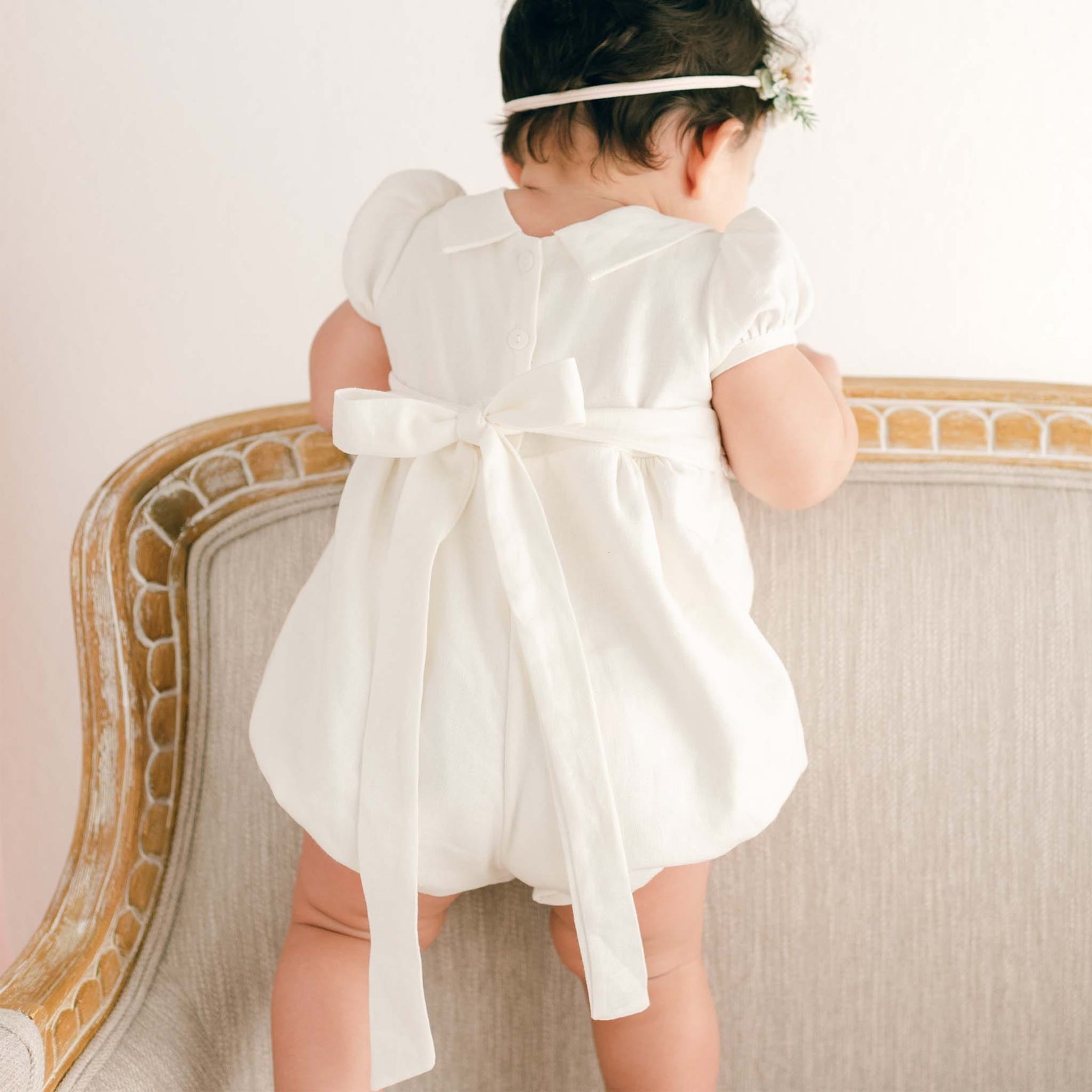 A baby in an Emma Christening Bubble Romper, coupled with a floral headband, stands on a beige armchair. Featuring a large bow and delicate ivory Venice lace accents, the little one reaches over the back of the chair, creating an endearing scene.