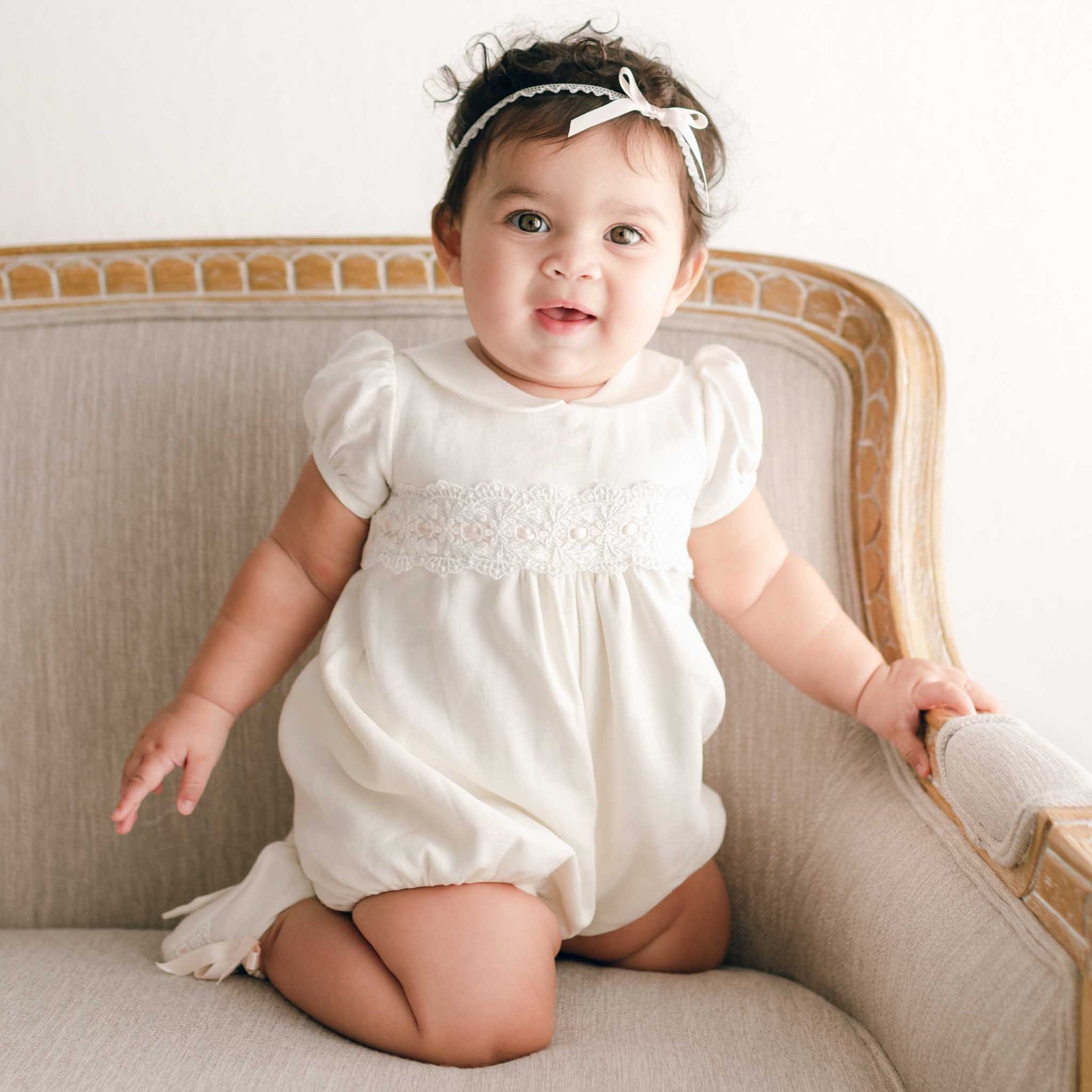A baby wearing an ivory Emma Christening Bubble Romper is seated on a beige armchair. With curly hair and a cheerful smile, the baby gazes towards the camera while the armchair showcases elegant wooden detailing along its edges.