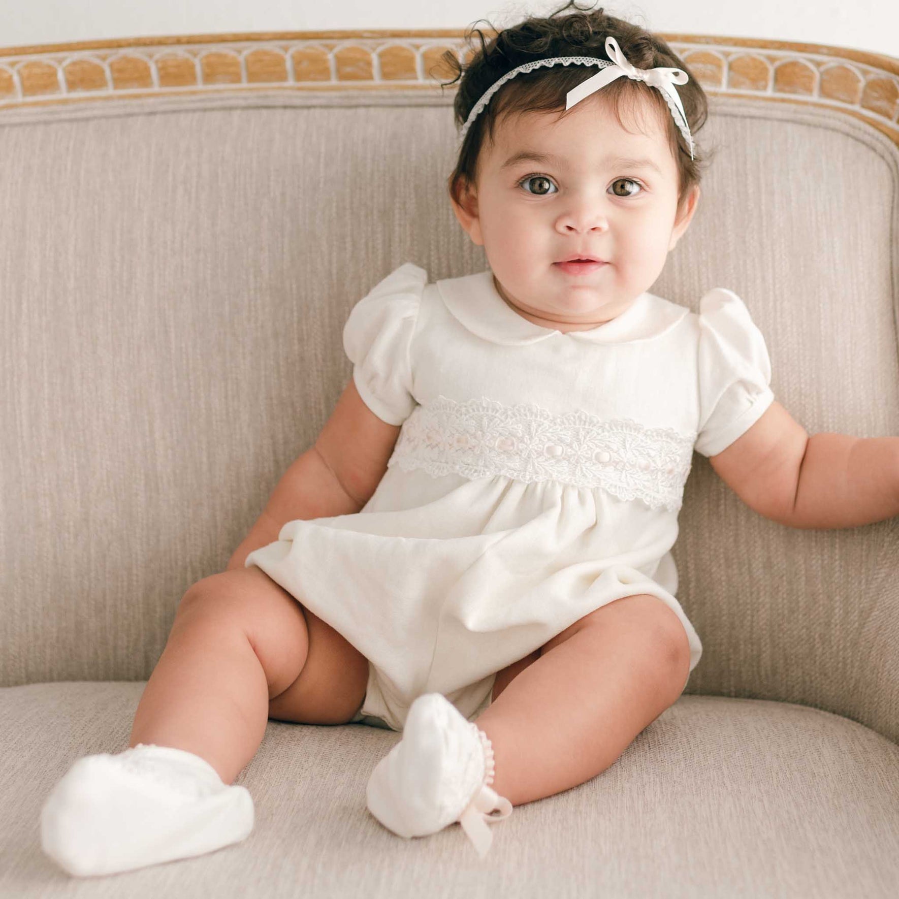 A baby, dressed in the Emma Christening Bubble Romper, sits on a beige cushioned chair. The white outfit is adorned with ivory Venice lace, and the little one wears socks and a headband with a bow, looking forward.