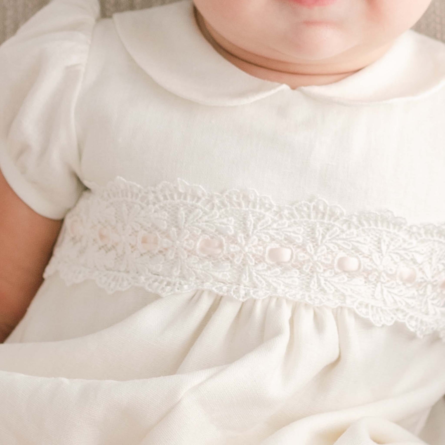 A close-up of a baby wearing the Emma Convertible Christening Gown Skirt & Romper Set, featuring a cream-colored design adorned with ivory lace and decorative trim across the chest. Handmade in the USA, this set includes a Peter Pan collar and puffed sleeves. The image perfectly highlights the baby's upper torso and neck.