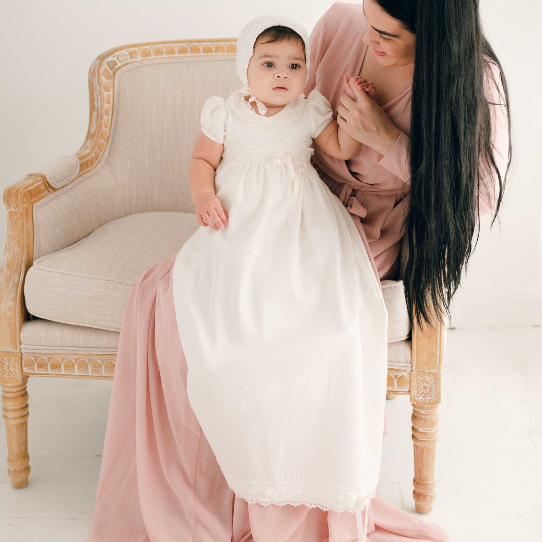 A woman in a pink dress sits on a light-colored chair, holding a baby dressed in the Emma Convertible Christening Gown Skirt & Romper Set, featuring an ivory lace design. The baby has a neutral expression, and both are in a softly lit room with a white background. Their attire, handmade in the USA, contributes to the serene ambiance.