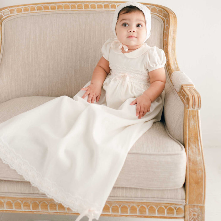 A toddler wearing the Emma Convertible Christening Gown Skirt & Romper Set, featuring a white dress with ivory lace details, sits on a beige cushioned armchair. The elegant design of the outfit harmonizes beautifully with the decorative wooden frame of the chair. With a neutral expression, the child looks upwards, embodying timeless charm.