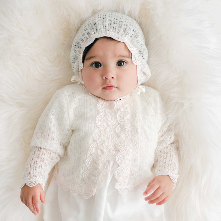 Baby wearing a white knit sweater and bonnet on a soft white background