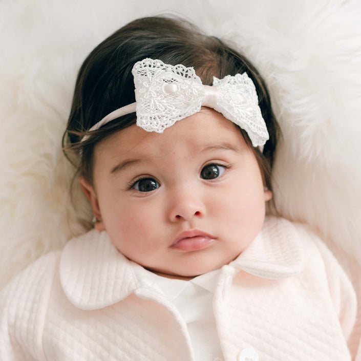 A baby lying on a white faux fur surface, wearing a pale pink quilted cardigan over an ivory dress and an ivory lace bow headband.