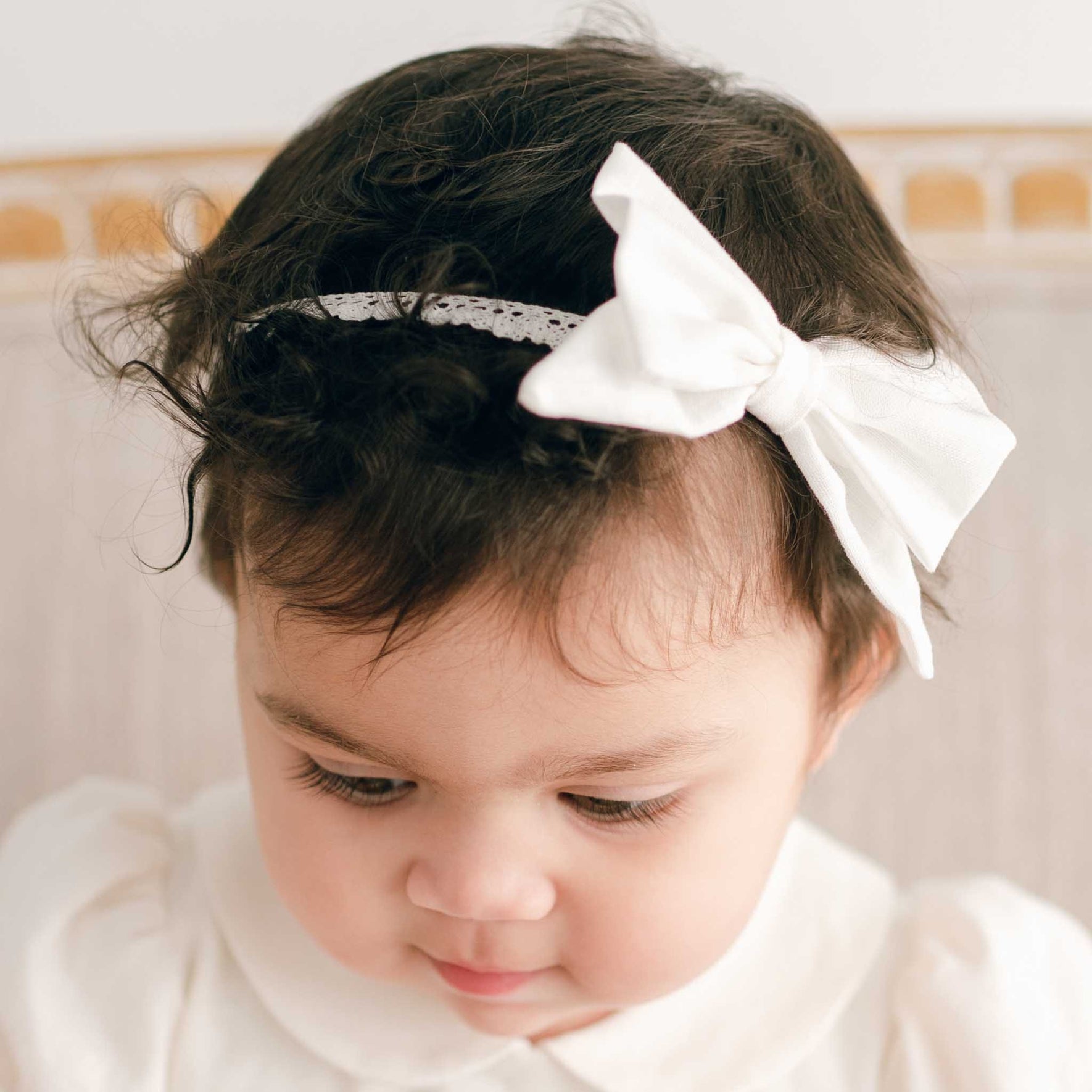 A young child with dark curly hair wears an Emma Linen Bow Christening Headband, beautifully complementing their white Christening outfit. The child looks down, with the softly blurred background enhancing the gentle elegance of the scene.