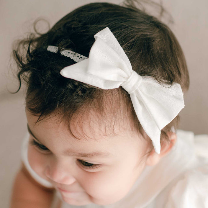 A close-up of a baby wearing the Emma Linen Bow Christening Headband as part of a charming outfit. The baby, with dark curly hair, gazes downward with a slight smile, emphasizing the adorable features accentuated by the light ivory linen blend headband.