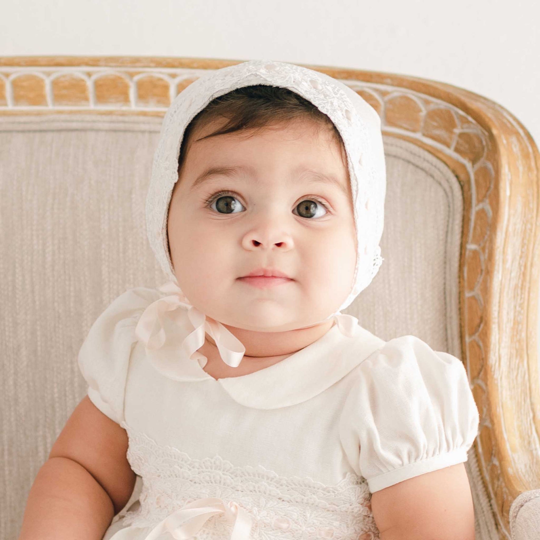 Front view of a baby girl wearing the Emma Christening Bonnet and matching dress, made from delicate ivory lace and tied with soft silk ribbons, seated on a beige upholstered chair and looking directly at the camera.