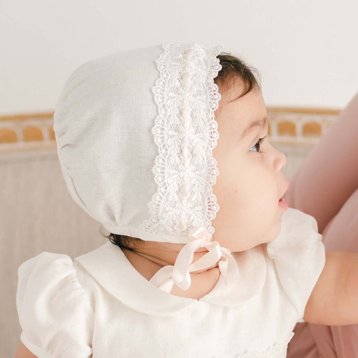 A close-up side view of a baby girl wearing the Emma Christening Bonnet and matching dress, looking thoughtfully to the right. This perspective features the intricate ivory Venice lace along the bonnet’s edge.