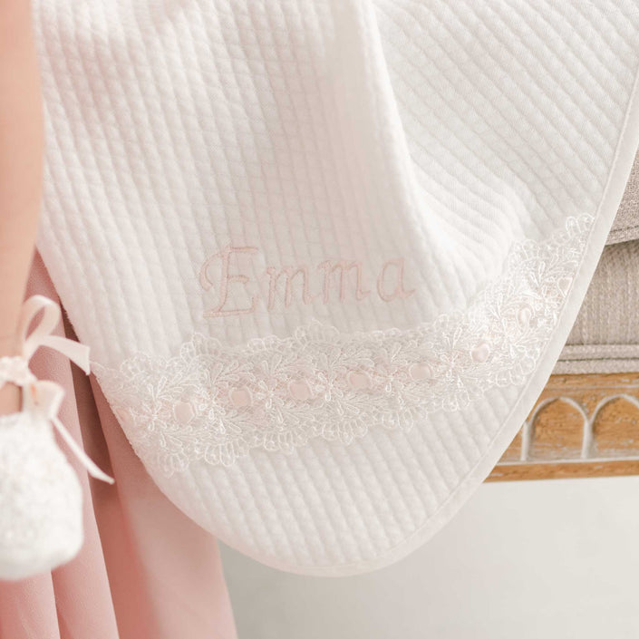 The Emma Personalized Blanket, a white quilted cotton piece, is elegantly draped over the edge with "Emma" embroidered in pink. It showcases a decorative lace trim with small pink embellishments—a beautiful complement to the white lace shoe adorned with a pink ribbon tie in the corner, making it ideal for a charming Christening blanket ensemble.