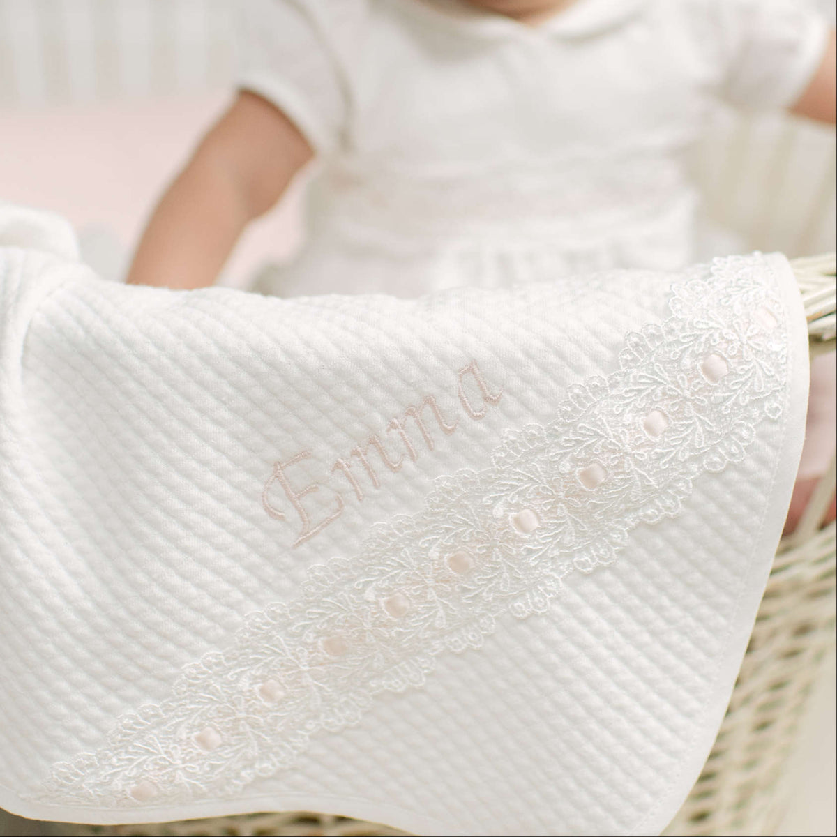 A close-up of an ivory textured baby blanket with delicate lace trim and the name 'Emma' embroidered in pale pink, draped over a baby in a wicker bassinet.