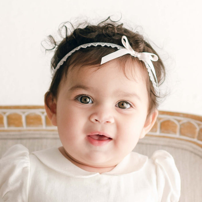 An adorable baby with curly hair and an Emma Pink Silk Ribbon Christening Headband sits on a chair. The little one is dressed in a white outfit, complete with a silk ribbon bow, gazing at the camera with a neutral expression. The background is plain and light-colored.